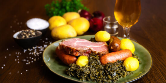 Ein klassisches norddeutsches Grünkohlgericht auf einem grün-glasierten Teller: zartes Kasseler, herzhafte Pinkel- und Kochwurst, goldgelbe Salzkartoffeln und frisch geschmorter Grünkohl. Im Hintergrund liegen Kartoffeln, rote Zwiebeln, grobes Salz, Pfefferkörner, ein Schnapsglas und ein frisch eingeschenktes Bier – warmes, rustikales Ambiente im Hotel & Restaurant am Huntepadd.